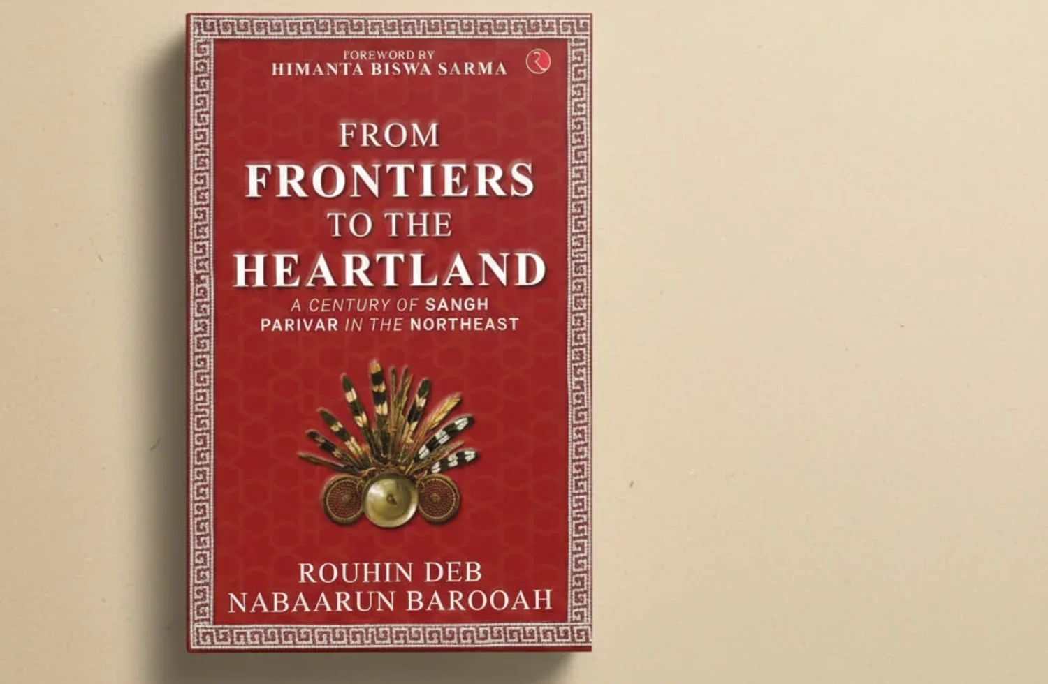 Book cover of From Frontier to the Heartland: A Century of Sangh Parivar in the Northeast by Rouhin Deb and Nabaarun Barooah, featuring a red patterned cover with a traditional Northeastern tribal ornament placed at the center, photographed against a minimal beige background.